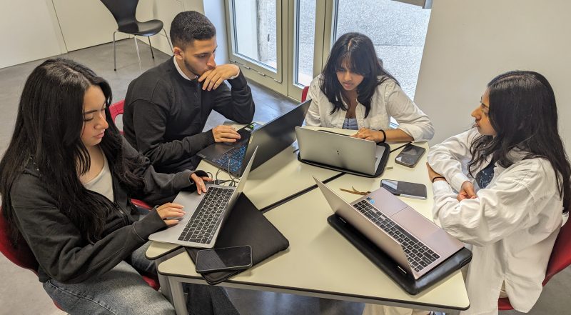four students sitting at a table working on laptops